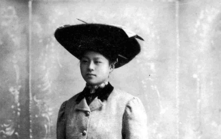 An early 20th century portrait of a Chinese woman wearing a long formal coat and a black hat.